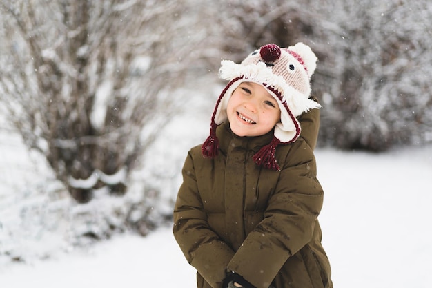 Il ragazzino sveglio in divertente cappello invernale cammina durante una nevicata all'aperto attività invernali per bambini