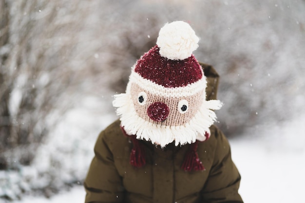 Il ragazzino sveglio in divertente cappello invernale cammina durante una nevicata all'aperto attività invernali per bambini