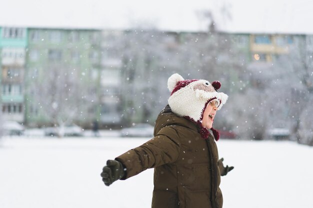 Il ragazzino sveglio in divertente cappello invernale cammina durante una nevicata all'aperto attività invernali per bambini