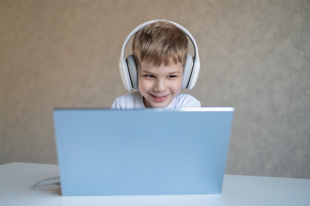 Il ragazzino sveglio sta guardando i cartoni animati o un film su un computer portatile Il bambino caucasico si siede a un tavolo a casa e ascolta la musica sulle cuffie