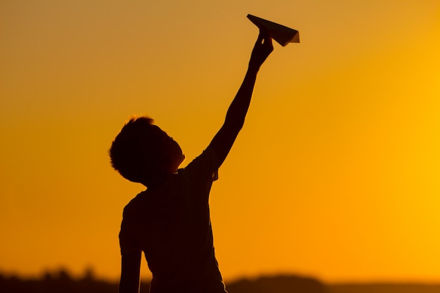 Il ragazzino tiene un aeroplano di carta in sua mano al tramonto. Un bambino alza la mano in cielo e gioca con gli origami la sera in strada