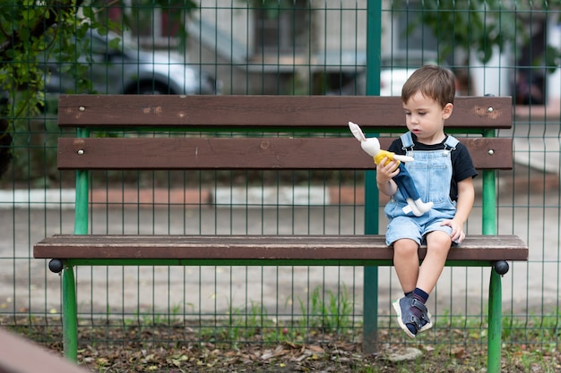 Il ragazzo adorabile si siede su una panchina nel parco giochi e guarda il coniglietto giocattolo