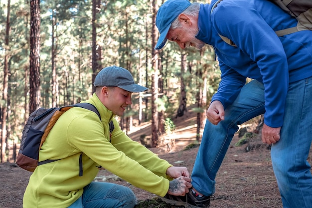 Il ragazzo aiuta suo nonno ad allacciarsi una scarpa mentre camminano insieme nella foresta di montagna godendosi la natura e la sana attività La nuova generazione aiuta quella più anziana L'avventura non ha età