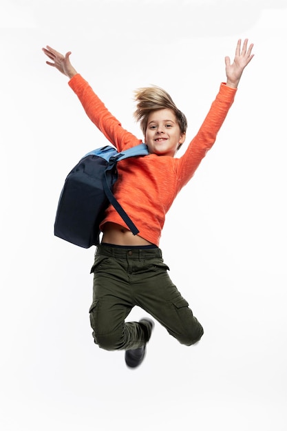 Il ragazzo allegro con uno zaino sta saltando. Un ragazzo in jeans scuri e un maglione arancione. Positività, attività e movimento. Di nuovo a scuola. Isolato su sfondo bianco. Verticale.