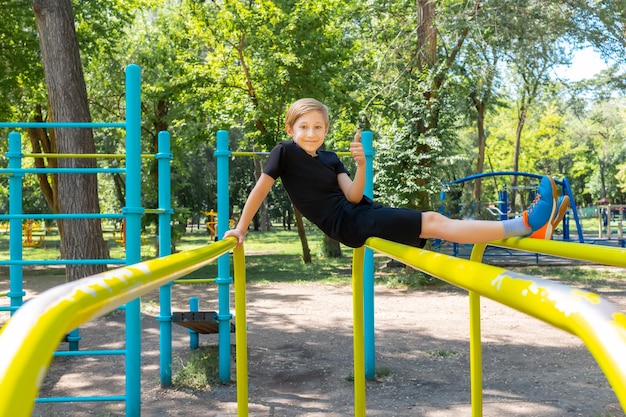 Il ragazzo allegro nel parco si siede sulle sbarre ginniche ride e mostra i pollici in su