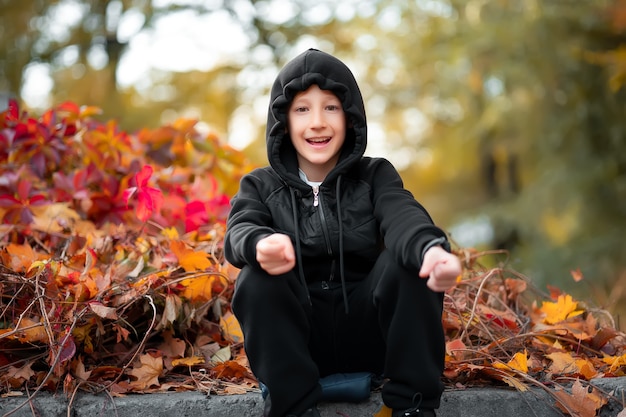 Il ragazzo allegro si siede sul marciapiede nel parco vicino al cespuglio rosso autunnale.