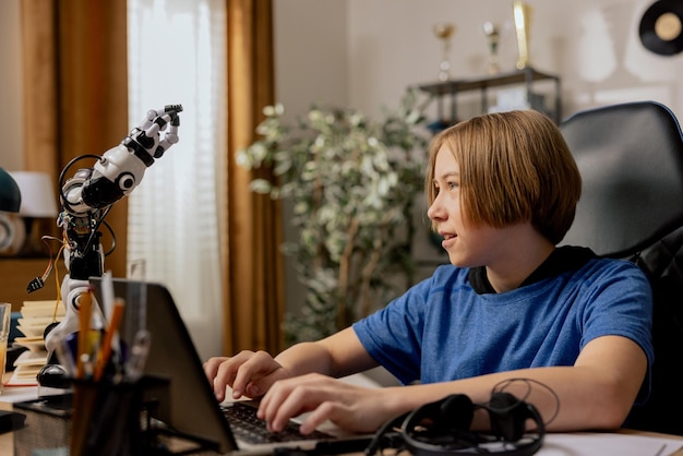 Il ragazzo ambizioso e sorridente intelligente si siede alla scrivania davanti al laptop guardando il robot che sta testando l'adolescente