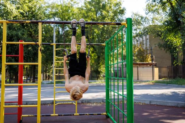 Il ragazzo appeso agli anelli sportivi a testa in giù tenendoli con le mani Street workout su una barra orizzontale nel parco della scuola