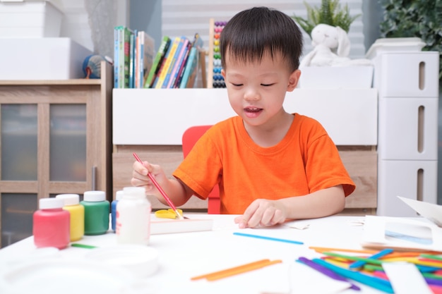 Il ragazzo asiatico del bambino gode di di usare la colla che fa le arti a casa