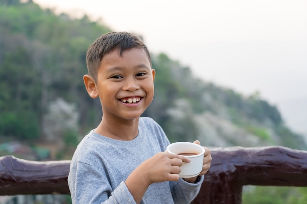 Il ragazzo asiatico del bambino sta bevendo l&#39;acqua dal vetro. Con lo sfondo delle montagne