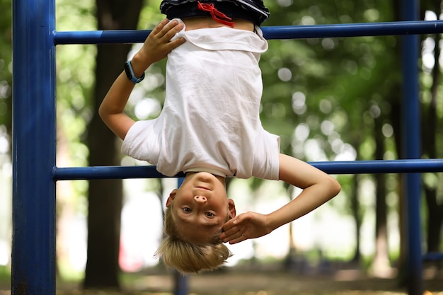 Il ragazzo atleta sul campo sportivo del parco ha preso i piedi sulle scale e appeso a testa in giù mostra diversi gesti con le mani