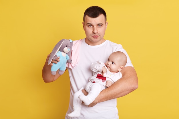 Il ragazzo attraente caucasico con l'espressione facciale seria che posa contro il muro giallo, indossa casualmente, tiene la neonata nelle mani, mostrando il coniglio di peluche al bambino, sembra stanco.