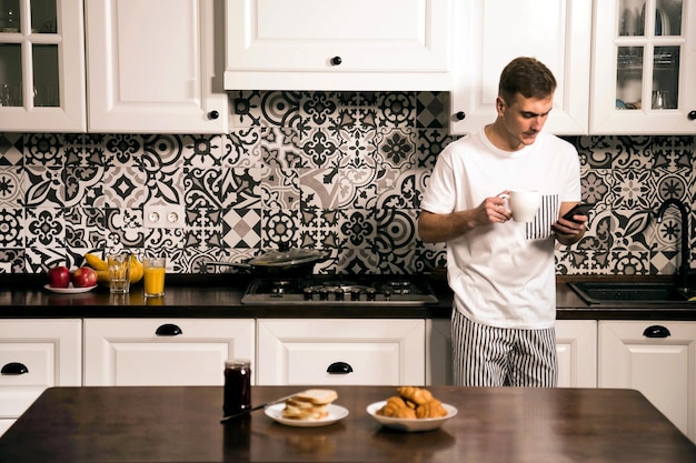 Il ragazzo barbuto attraente legge i messaggi sullo smartphone in piedi in cucina