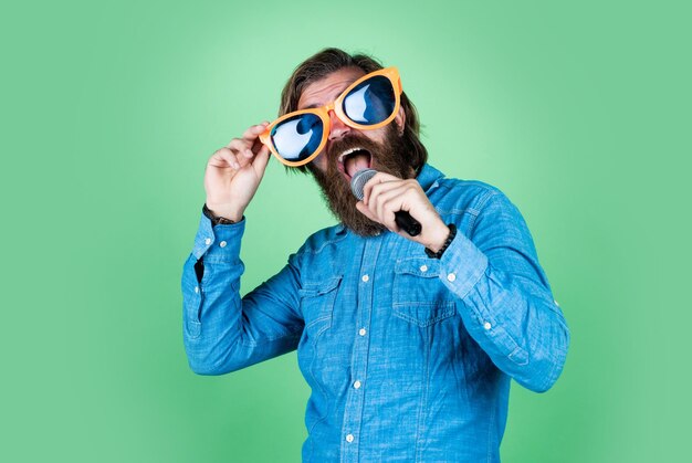 Il ragazzo barbuto maturo ha i capelli lunghi che indossa occhiali da festa e canta divertendosi con il microfono