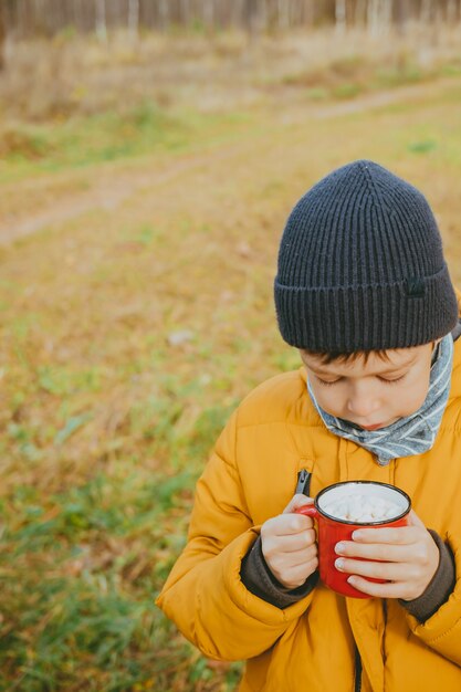 Il ragazzo beve cioccolata calda dalle tazze nella foresta il ragazzo con una giacca gialla tiene in mano cioccolata calda calda