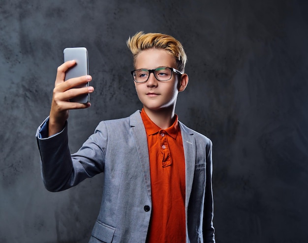 Il ragazzo biondo dell'adolescente in una giacca e occhiali da vista tiene lo smartphone.