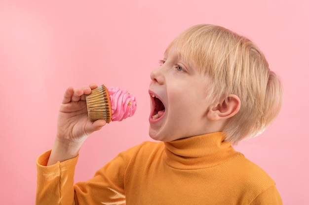 Il ragazzo biondo mangia il cupcake con la glassa rosa. Il bambino vuole mordere un pezzo di muffin.