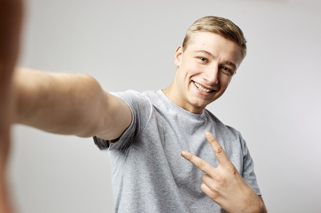 Il ragazzo biondo sorridente vestito con una maglietta bianca mostra v cantare e fa un salfie