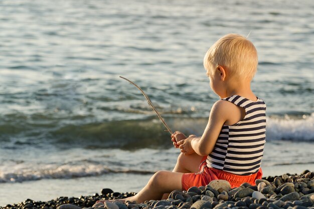 Il ragazzo biondo sveglio in camicia a strisce si siede su Pebble Beach e tiene la canna da pesca improvvisata.