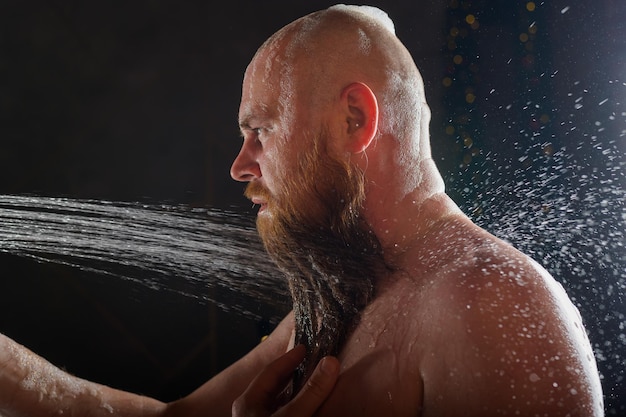 Il ragazzo calvo fa la doccia Un uomo brutale con la barba rossa è in piedi in bagno sotto un getto d'acqua e si lava Spray sparso su uno sfondo nero