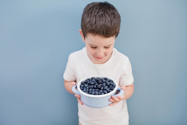 Il ragazzo carino del bambino tiene il mirtillo fresco in una pentola blu Bambino con le bacche su sfondo blu