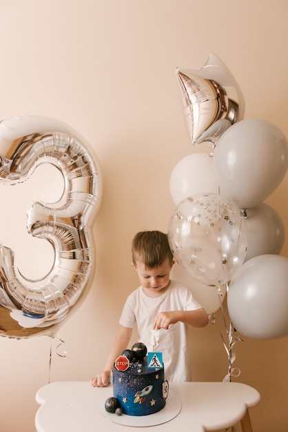 Il ragazzo carino di 3 anni festeggia il suo compleanno e mangia una deliziosa torta bellissima, foto di un bambino con palloncini