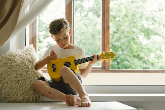 Il ragazzo carino impara a suonare la chitarra Ukulele gialla sul davanzale vicino alla finestra Casa accogliente Stile di vita per le vacanze estive