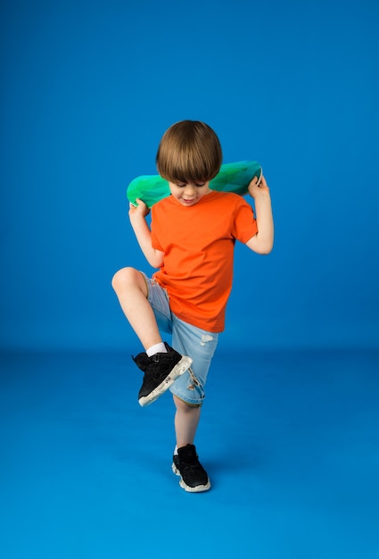 Il ragazzo che balla con i capelli castani in una maglietta arancione e pantaloncini di jeans tiene uno skateboard su una superficie blu con spazio per il testo