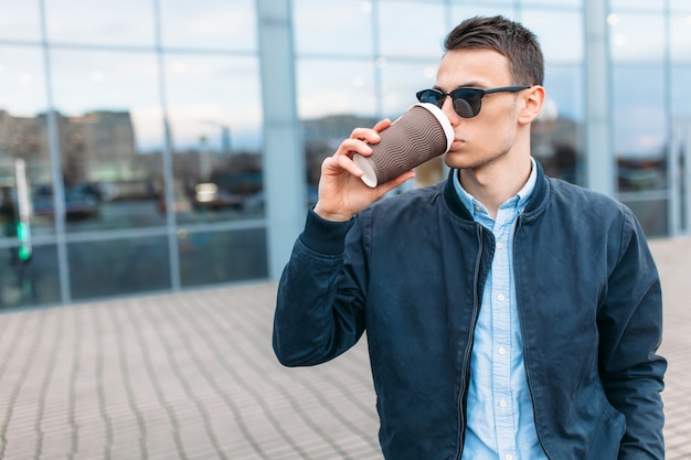 Il ragazzo con gli occhiali da sole alla moda, un uomo cammina per la città e beve il caffè da una tazza di carta, un bel ragazzo cammina e riposa, il ragazzo sullo sfondo di un edificio moderno, ufficio