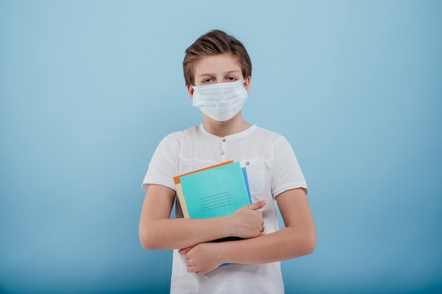 Il ragazzo con la maschera medica ha quaderni e libri in mano isolati su sfondo blu