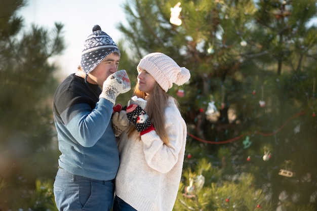 Il ragazzo con la ragazza cammina e bacia nella foresta invernale con una tazza di bevanda calda. Un'accogliente passeggiata invernale attraverso i boschi con una bevanda calda. Coppia di innamorati, vacanze invernali