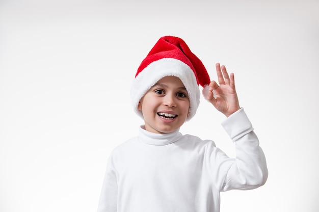 Il ragazzo con un cappello rosso da Babbo Natale sorride e mostra un gesto di "ok". Concetto di natale.