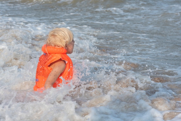 Il ragazzo dai capelli chiari in un giubbotto gonfiabile arancione sta nuotando nel mare.