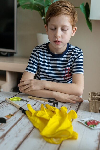 Il ragazzo dai capelli rossi del bambino del giardinaggio domestico in una maglietta a righe sta piantando piantine stagionali in vasi ecologici