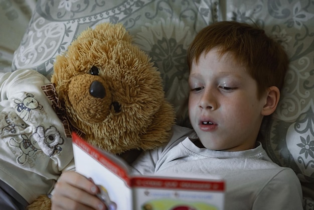 Il ragazzo dai capelli rossi del bambino legge un libro prima di andare a letto si trova a letto con un ritratto del primo piano dell'orsacchiotto a casa