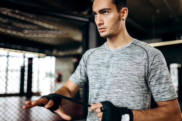 Il ragazzo dai capelli scuri vestito con una maglietta grigia e pantaloncini neri si trova in palestra accanto al ring e avvolge una benda sulla mano.