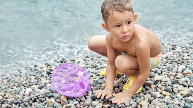Il ragazzo del bambino in età prescolare gioca con la rete di atterraggio viola raccogliendo ciottoli sulla spiaggia vicino all'acqua