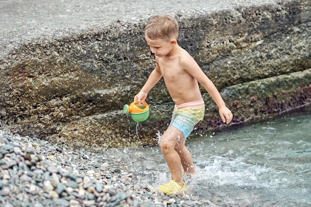 Il ragazzo del bambino in età prescolare porta l'annaffiatoio giocattolo sulla spiaggia di ciottoli vicino al molo