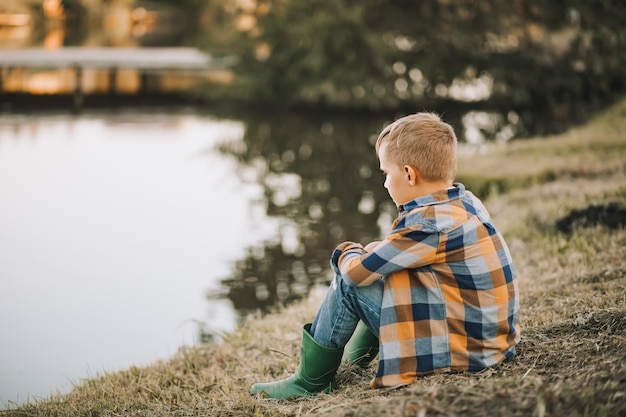 Il ragazzo del bambino sognante di pensiero triste si siede vicino a un fiume o un lago