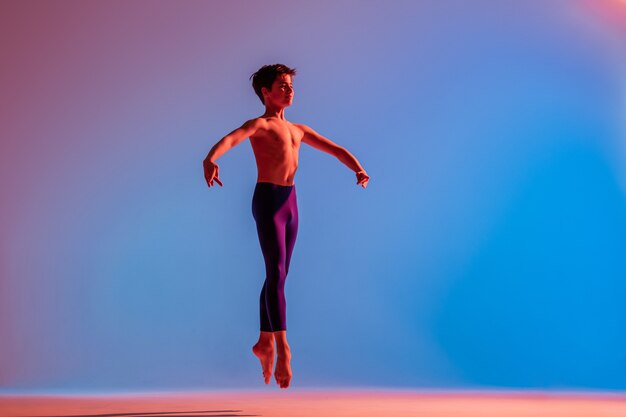 Il ragazzo di balletto teenager snello salta a piedi nudi sotto la luce colorata.