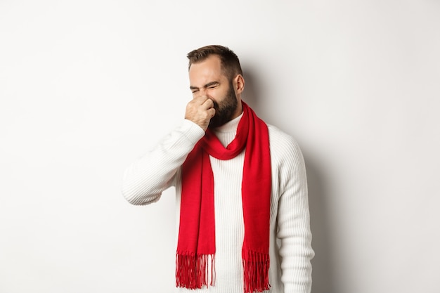 Il ragazzo dispiaciuto si chiuse il naso per il cattivo odore, facendo una smorfia disgustato, in piedi su sfondo bianco in maglione bianco con sciarpa rossa