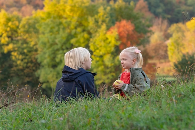 Il ragazzo e la ragazza biondi si siedono sull'erba e mangiano le mele Fratellini che riposano nella natura Bambini al picnic il giorno d'autunno