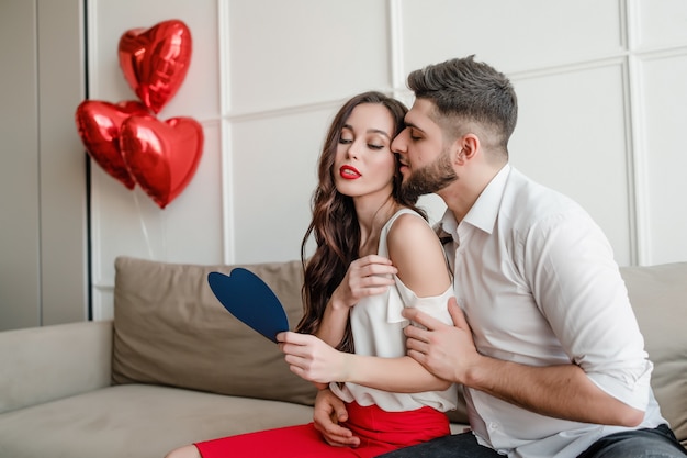 Il ragazzo e la ragazza con la carta blu del biglietto di S. Valentino e il cuore rosso hanno modellato i palloni che si siedono a casa sullo strato