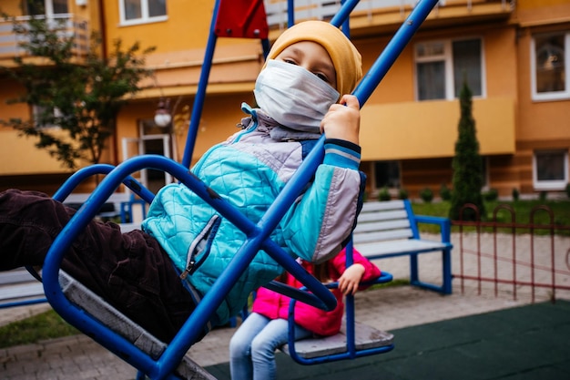 Il ragazzo e la ragazza della scuola elementare indossano maschere mediche e oscillano su un'altalena I bambini seri riposano nel parco giochi in autunno