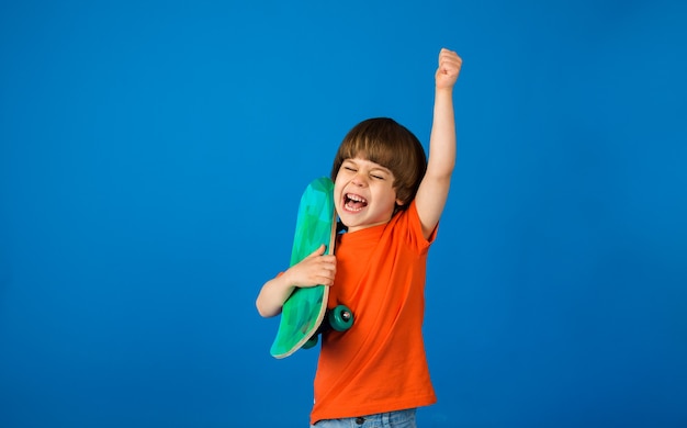 Il ragazzo felice del bambino in maglietta arancione tiene uno skateboard su una superficie blu con una copia dello spazio