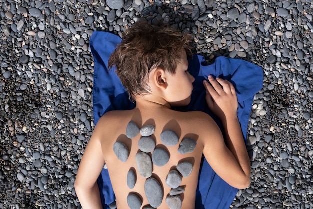 Il ragazzo giace su una spiaggia di ciottoli. Terapia della pietra. Relax, salute mentale dei bambini. Relax. Sole.