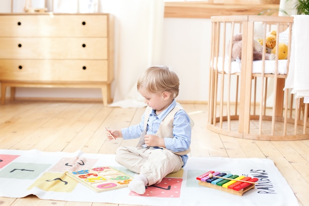 Il ragazzo gioca con i giocattoli di legno a casa. Giocattoli educativi in legno per il bambino. Ritratto di un ragazzo seduto sul pavimento nella stanza dei bambini in stile scandinavo. Giocattoli ecologici, arredamento per la camera dei bambini