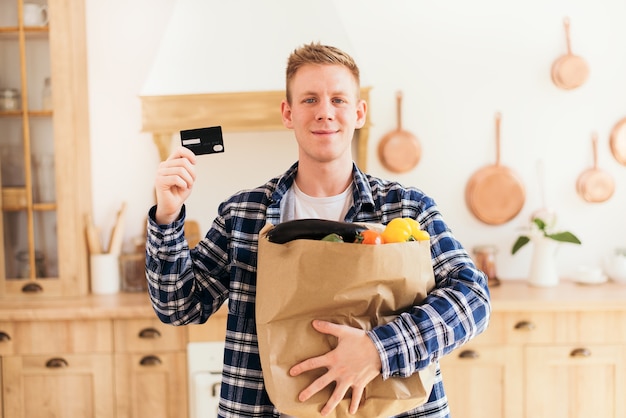 Il ragazzo ha in mano un pacco di generi alimentari e una carta di credito a casa in cucina shopping online e pagamento con carta