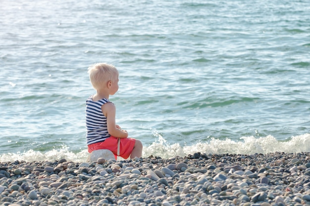 Il ragazzo in maglia a strisce si siede su una roccia in riva al mare.