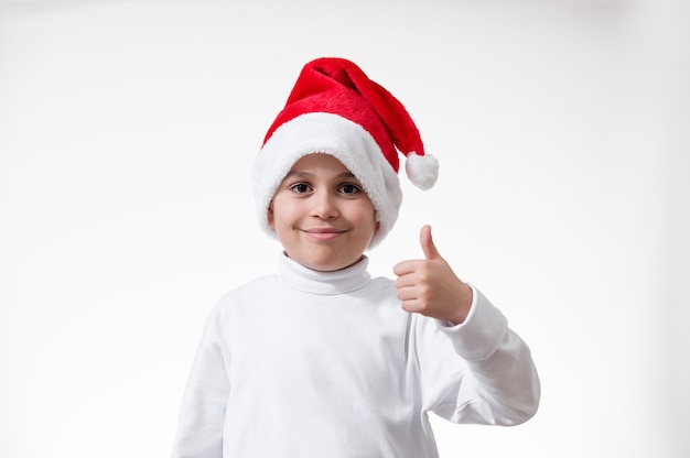 Il ragazzo in un cappello rosso della Santa sorride e mostra il suo pollice. Concetto di natale.